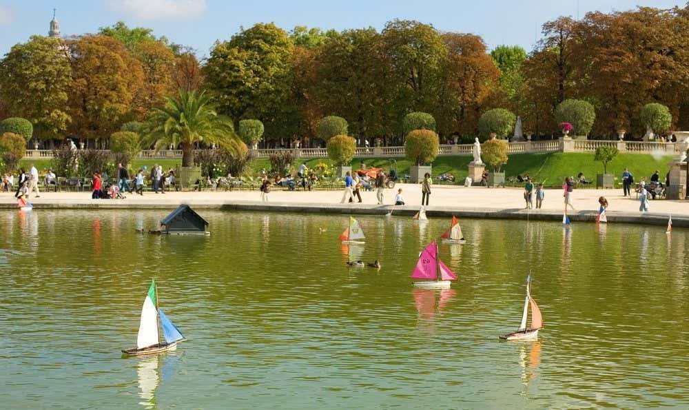 Bassin d'eau avec des bateaux miniatures flottants et plusieurs personnes se baladant dans le parc en arrière-plan