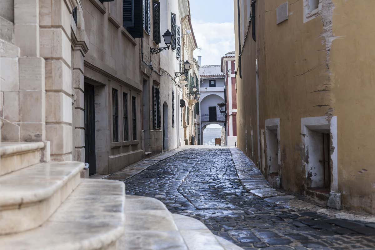 A narrow cobbled street in Mahón, with a wooden sign visible on the road.