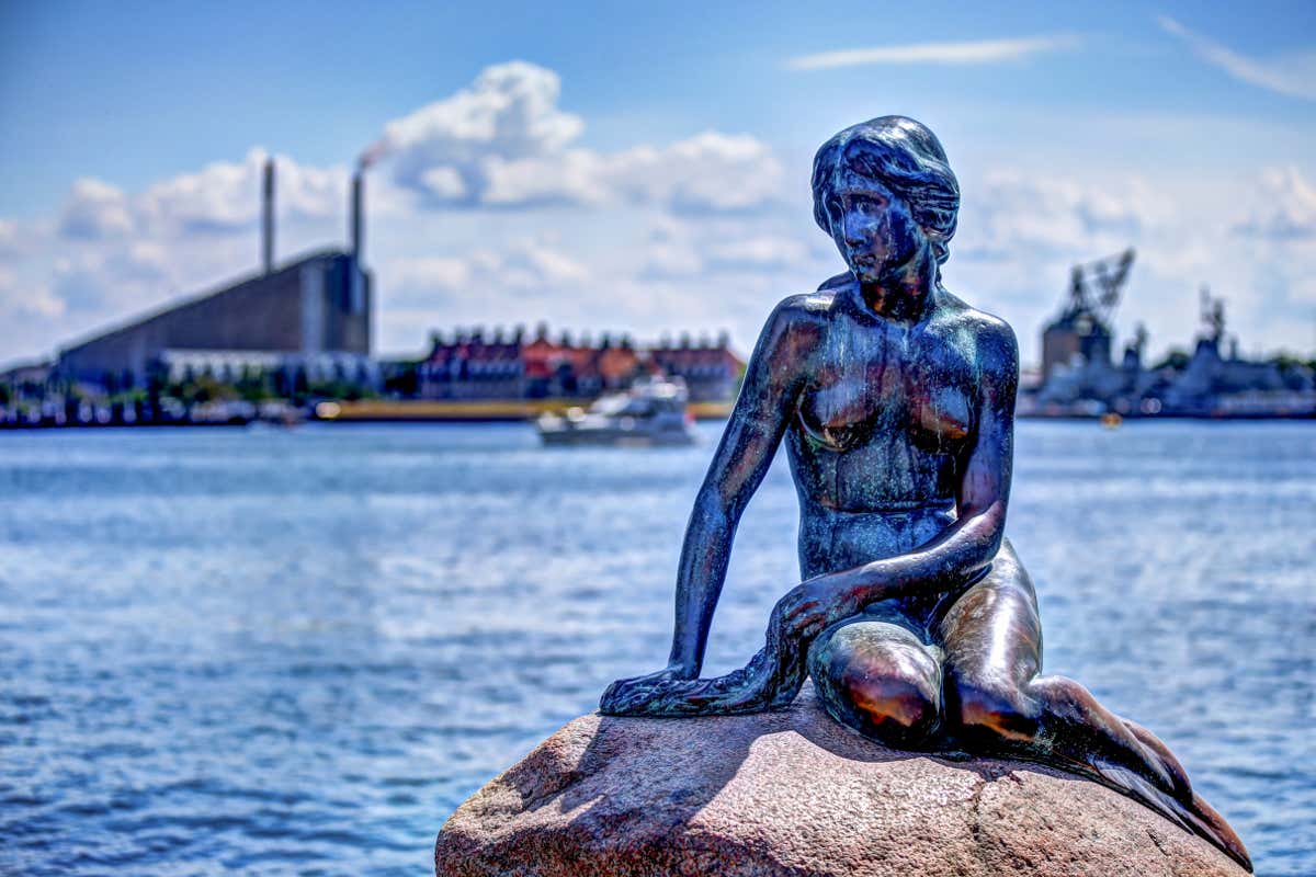 A metal statue of a mermaid on a rock by a wide open river in Copenhagen.