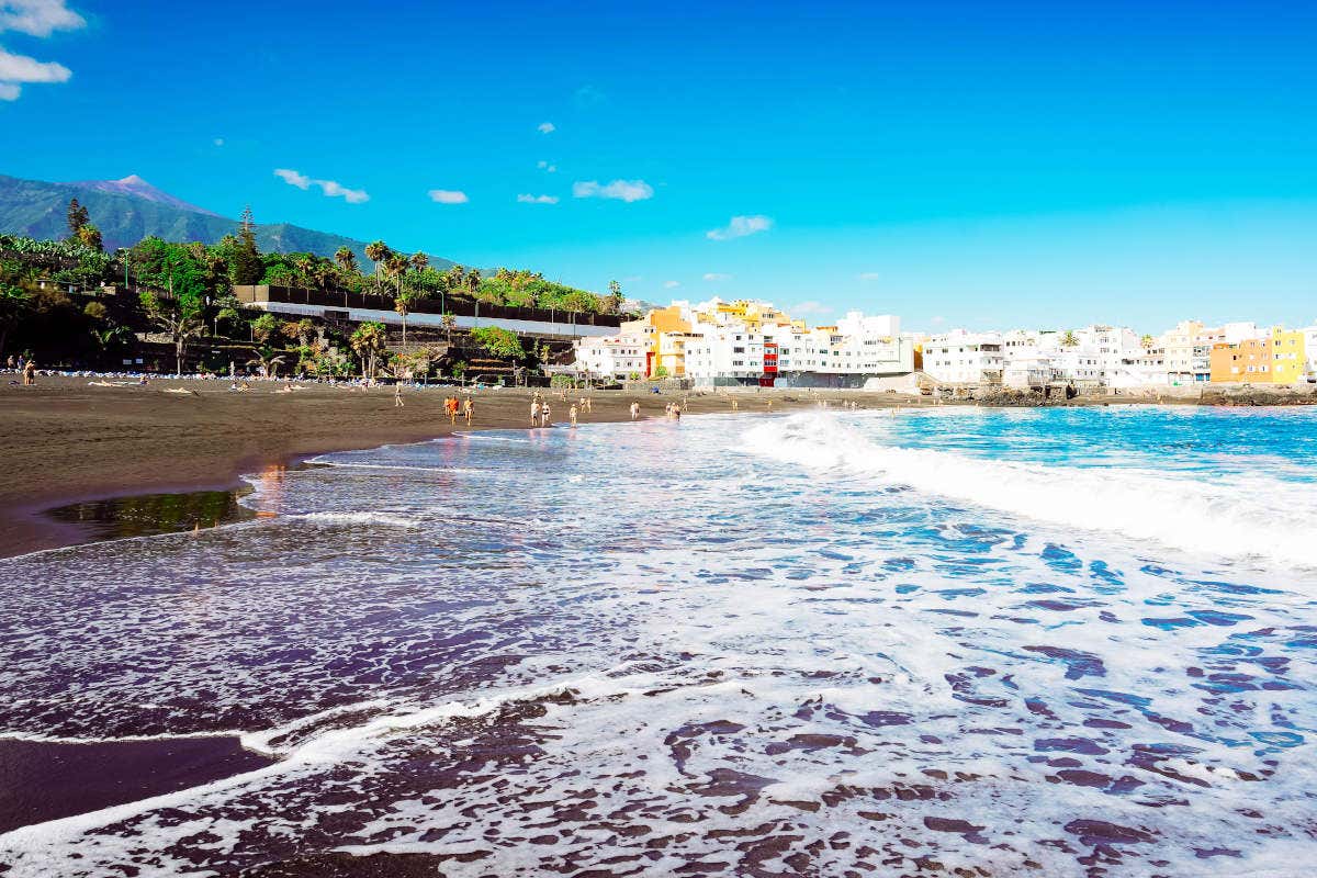 A Tenerife beach with black sand and a few people bathing on a clear day, with the silhouette of Mount Teide on the horizon.