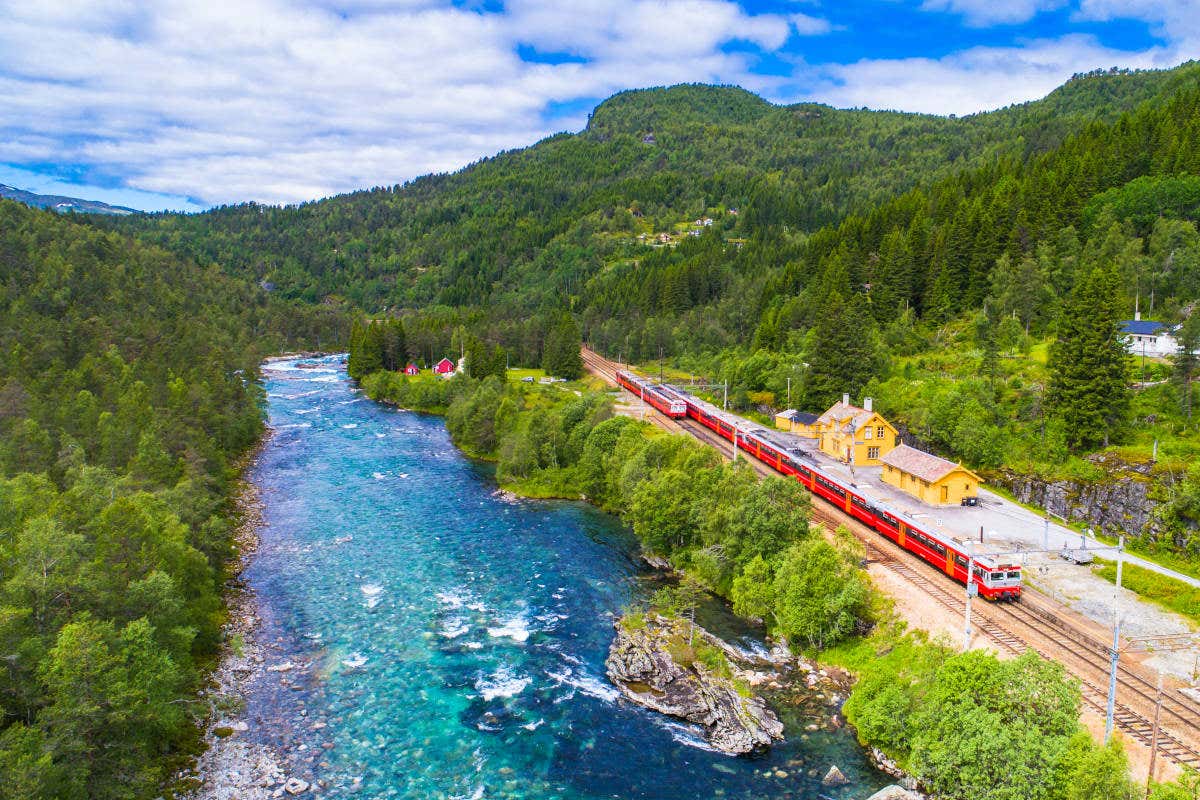 Train rouge à l'arrêt à une gare au bord d'une rivière et entourée de verdure