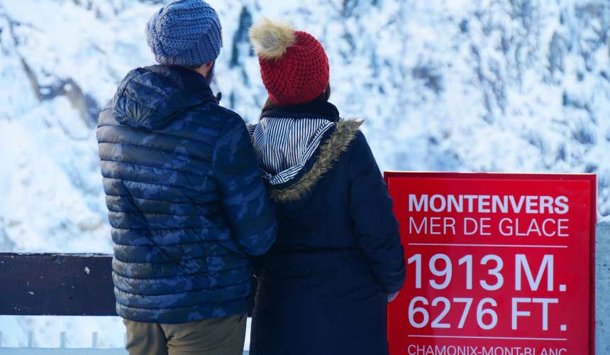 Due persone, una col giubbotto mimetico blu e cappello azzurro e l'altra con cappello rosso con pon-pon peloso e giubbotto blu navy, accanto a un cartello rosso con scritte bianche, ammirano il panorama del ghiacciaio Mer de Glace, nei pressi di Chamonix