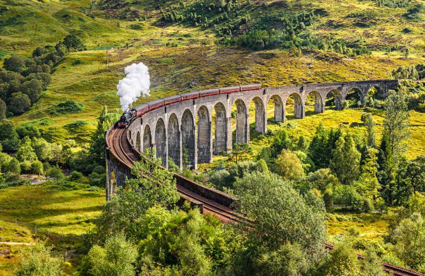 A red and black steam train runs through the mountains and Glenfinnan Viaduct in Scotland.