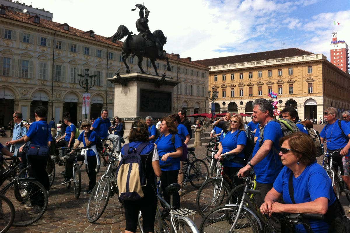 Un numeroso gruppo di persone con una maglietta blu in sella alle biciclette nella piazza di una città, pronti per un tour ecosostenibile che non inquina.