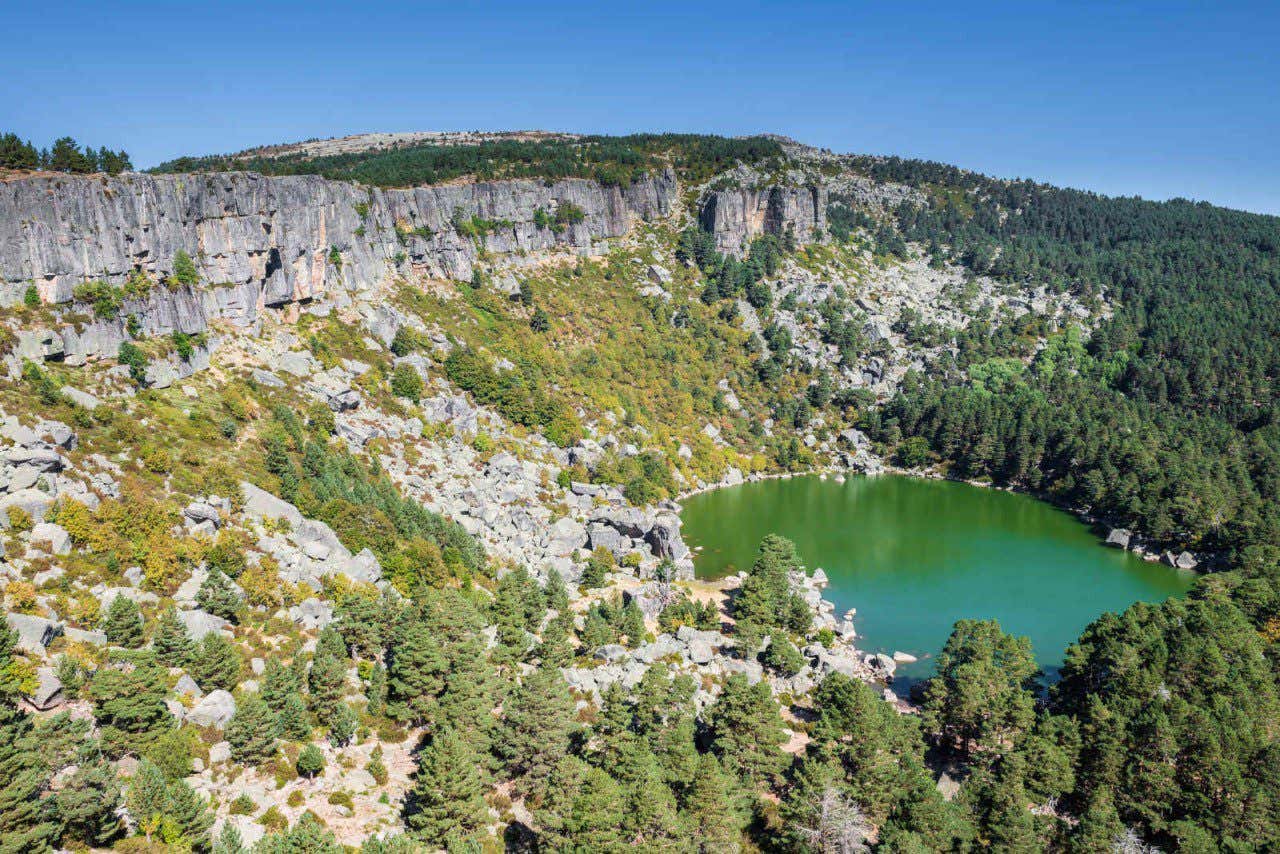 Vista panorámica de la laguna Negra de Soria en la parte baja de unos altos acantilados con vegetación arbórea 