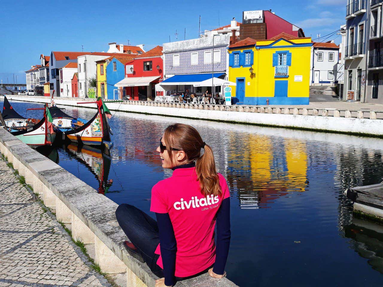 Anna Bondareva con una camiseta rosa con el logo de Civitatis fotografiándose frente a las casas coloridas y los barcos de los canales de Aveiro.