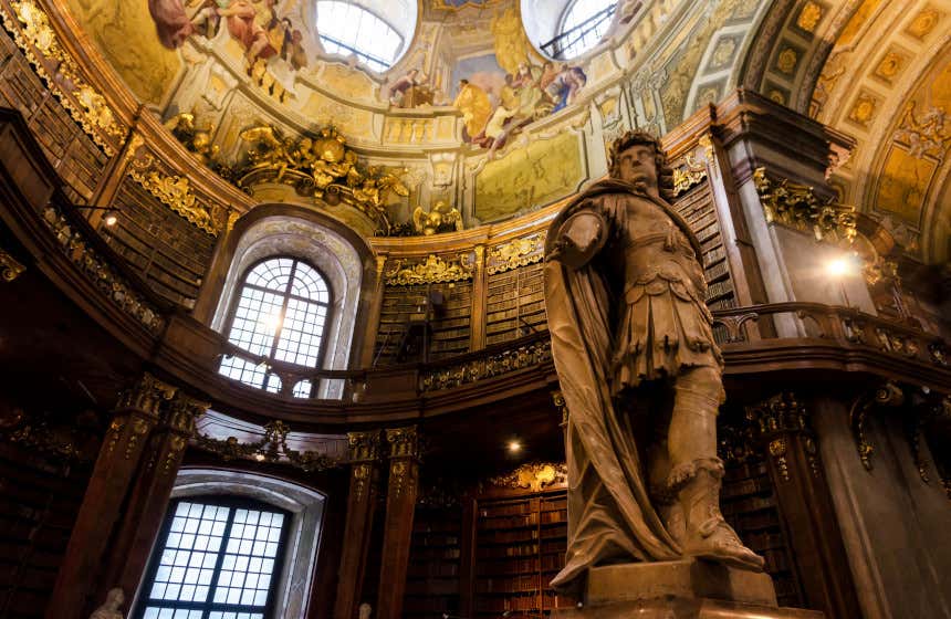 Entrada de la Biblioteca Nacional de Austria con una gran escultura de mármol en primer plano y, al fondo, varias estanterías de libros bajo una cúpula con coloridos frescos.