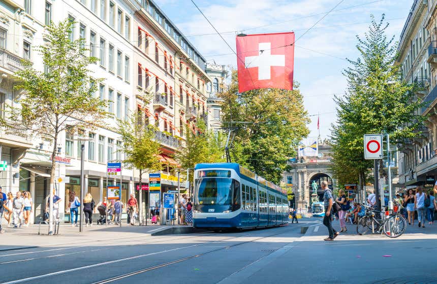 Bahnhofstrasse, una de las calles comerciales más importantes del centro de Zúrich, con gente paseando y un tranvía pasando bajo una bandera de Suiza.