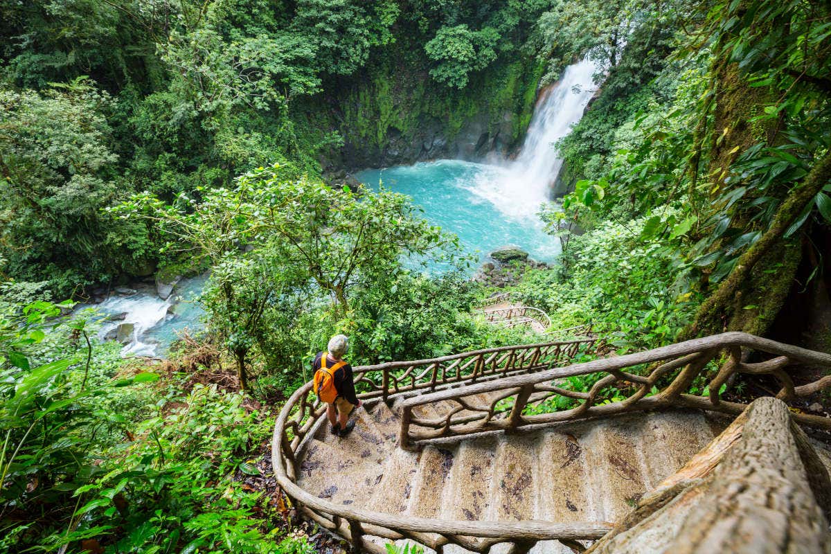 Vista panoramica su una maestosa cascata nella foresta della Costa Rica, con un turista che scende una scalinata verso la cascata, con lo zaino in spalla