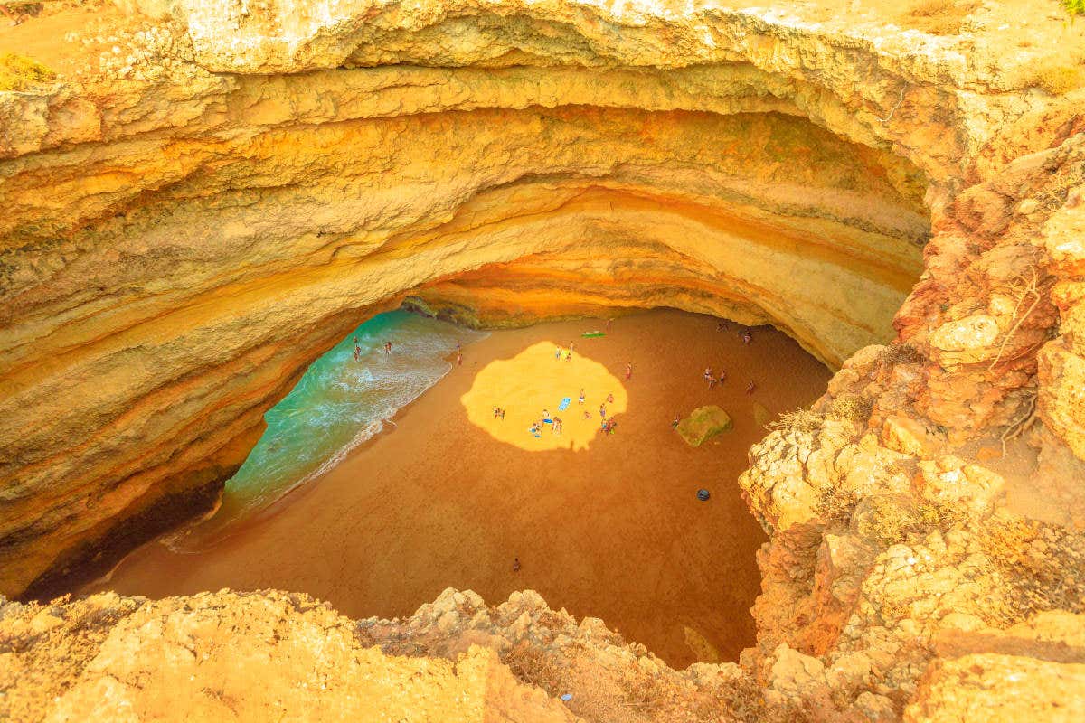 Aerial view looking down a skylight onto a beach with a small group of people surrounded by towering orange and yellow layered rocks