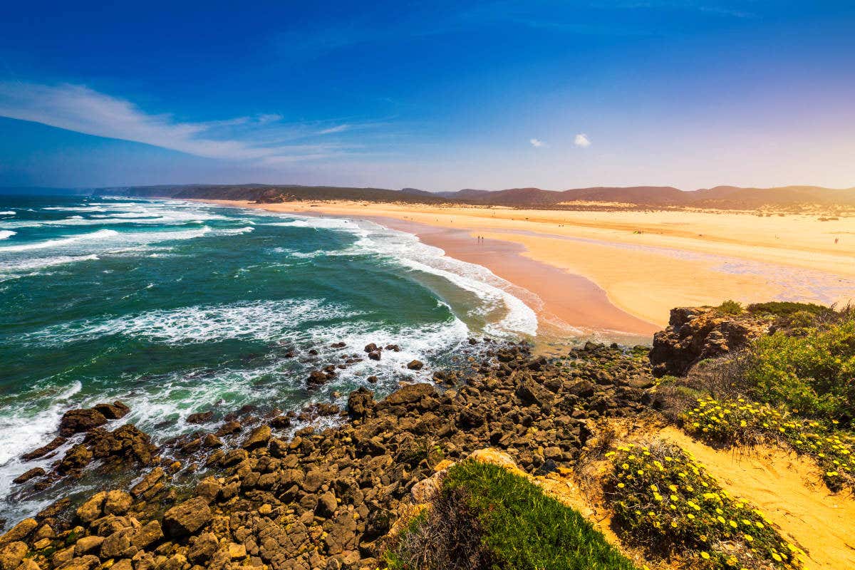 A long beach with fine golden sand and ocean waves crashing onto shore, a rocky area in the foreground, showing one of the best beaches in the algarve