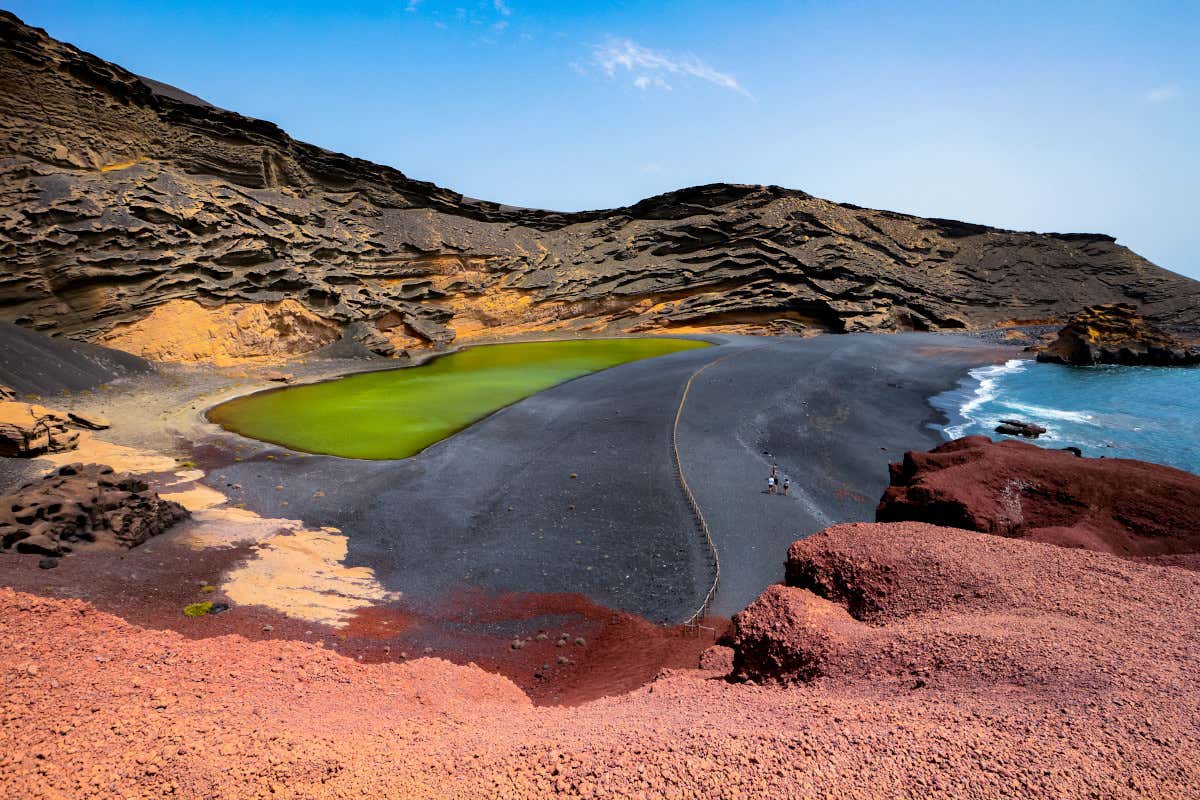 El Charco de los Clicos, un paisaje casi extraterrestre en la isla de Lanzarote donde se observan montañas rojizas de origen volcánico, una laguna de color verde y el propio mar azul en una playa de arena negra.