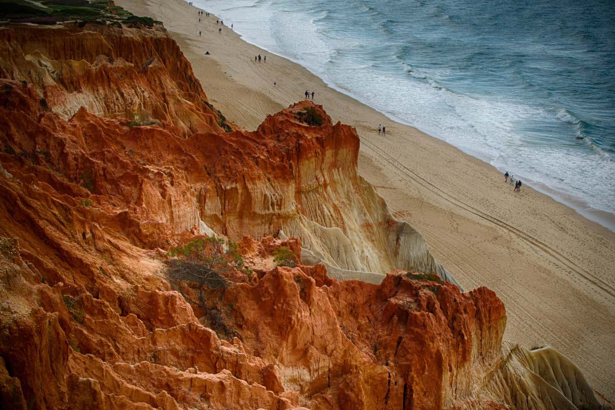 Red sanstone cliffs rise high above the sandy beach below with people walking along the sand next to the water, ocean waves crashing on the shore