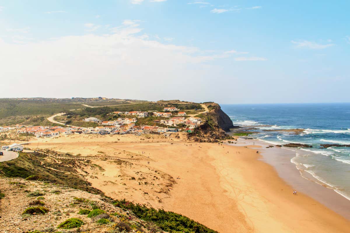 A golden sandy beach with a small beach town in the distance surrounded by lush green vegetation, ocean waves crashing onto the shore