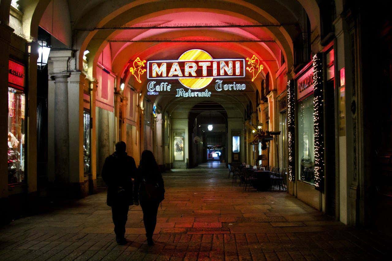 Due persone camminano di notte nei portici di Torino sotto la scritta "Martini" illuminata