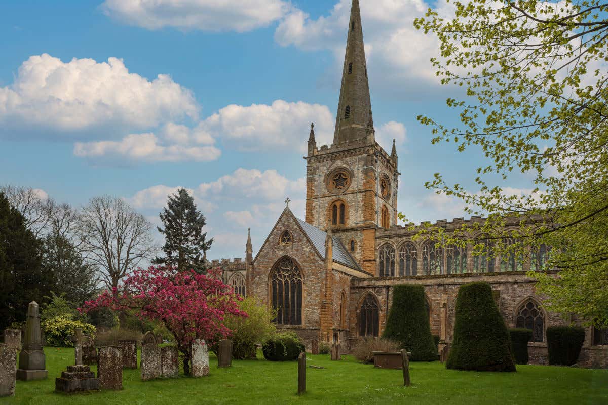 La iglesia de la Santísima Trinidad, ubicada en Stratford-upon-Avon. Vista de su torre y su fachada exterior frente a un antiguo cementerio adosado con varias tumbas sobre el césped, donde se encuentra una de las 10 tumbas más famosas del mundo