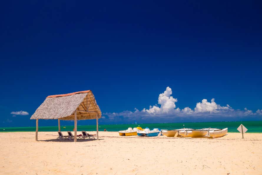 Cielo blu intenso con nuvole bianche in basso a destra sulla spiaggia di Santa Lucía, tra le migliori spiagge di Cuba, con un pedalò giallo e bianco e uno blu e bianco, due barche bianche e un cartello romboidale sulla destra e una costruzione in paglia e legno sulla sinistra con sotto tre sdraio