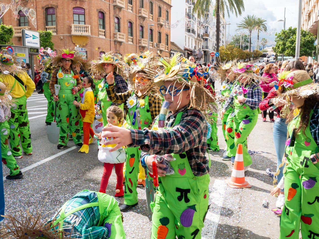 People dressed up as scarecrows walk along during a street parade featuring many children in bright green overalls on a sunny day in Cadiz