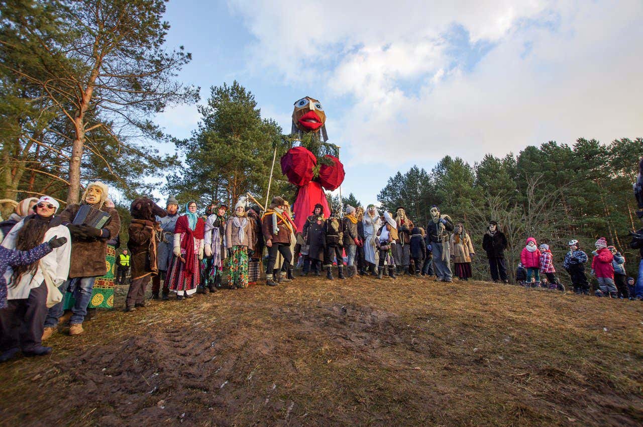 A field with a towering effigy featuring huge eyes and mouth dressed in red stands behind group of people also dressed up in masks standing in a field