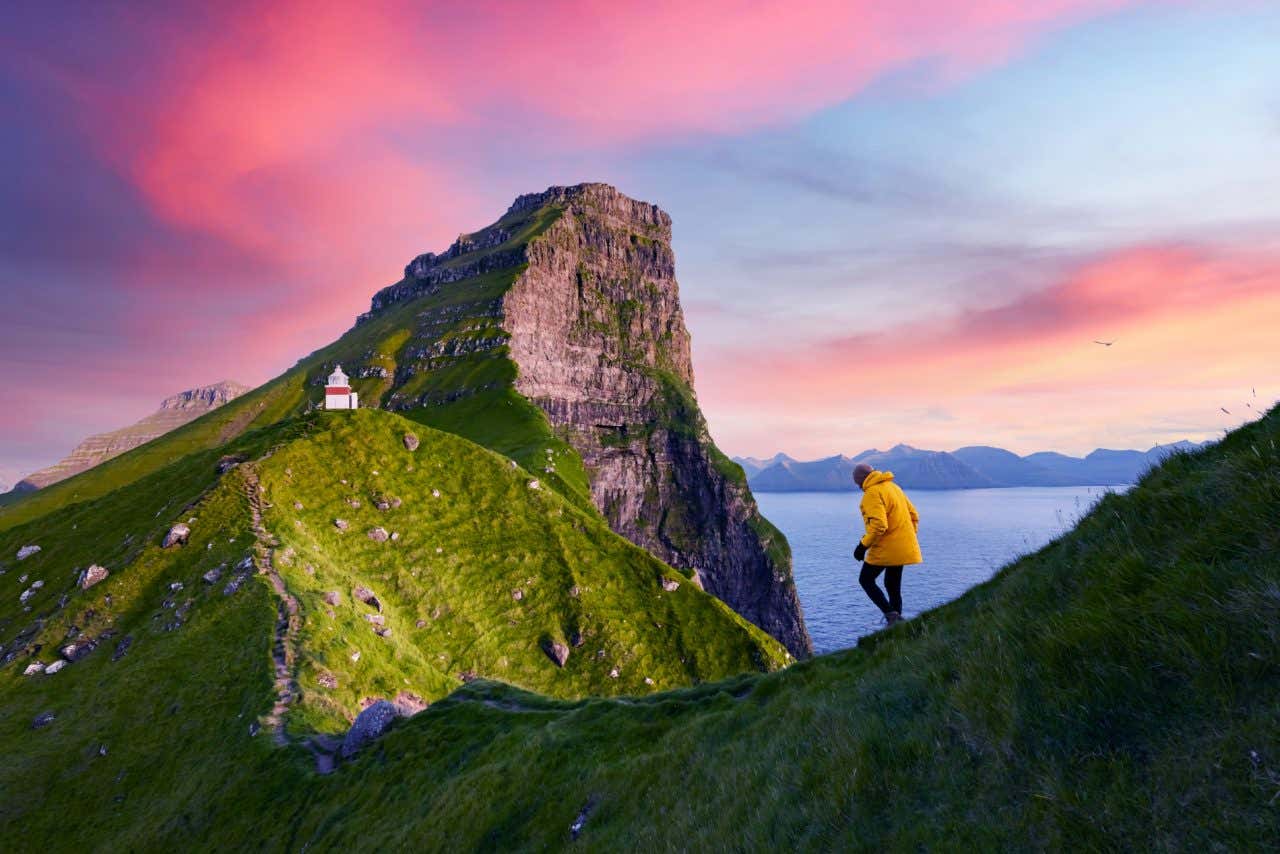 Un esploratore cammina lungo uno stretto sentiero sull'Isola di Kalsoy, con un piccolo faro bianco, il mare e le montagne sullo sfondo e il cielo al tramonto