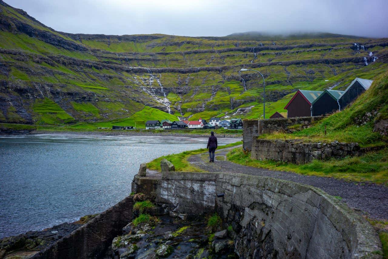 Il Villaggio di Tjørnuvík sulle rive del mare, con un una persona che cammina su un sentiero accanto alla scogliera, e alcune case ai piedi di un'altura verde