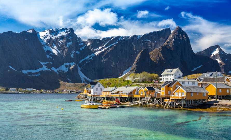 Traditional cabins or rorbu on Vestvågøy Island in the Lofoten Islands, Norway. Clear turquoise sea with snowy mountains surrounding the village.