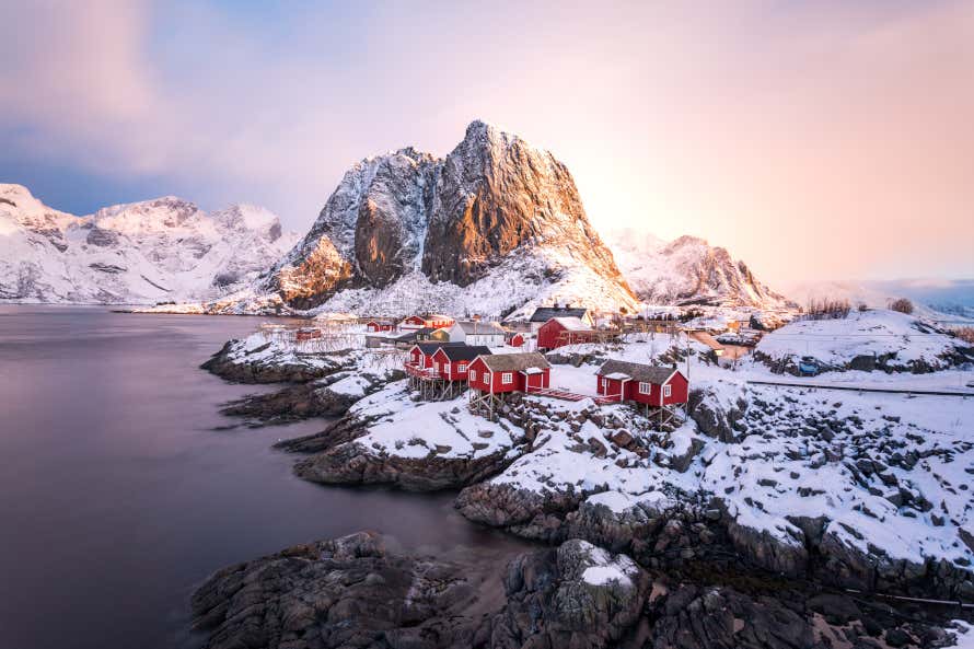 The Lofoten Islands in Norway in winter. A small village with red houses on a rocky island covered in snow with snow-capped mountains in the background.