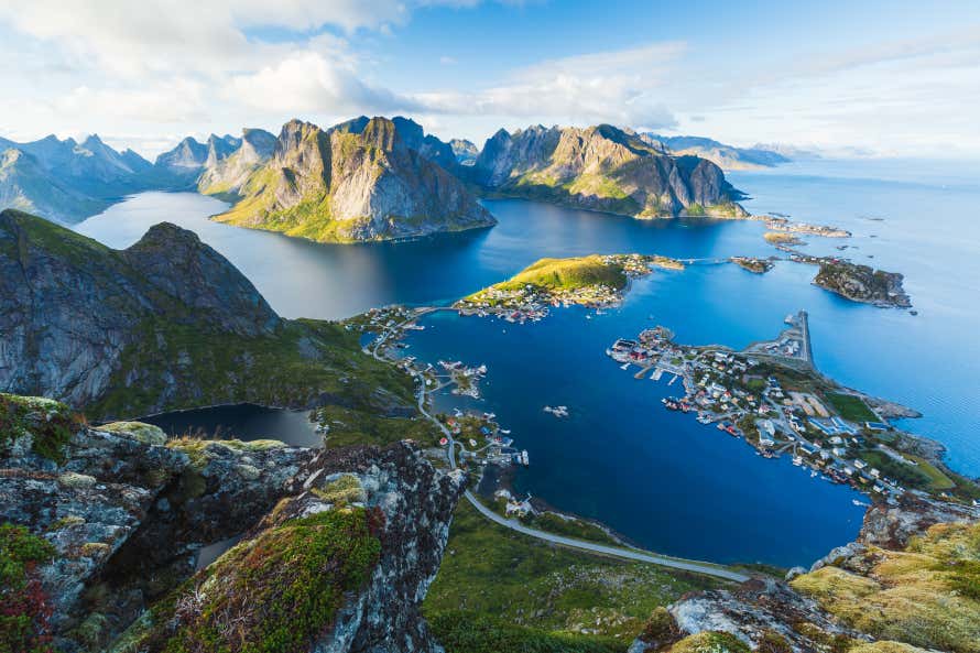 Panoramic photo of mountains jutting out of a calm sea with a fishing village tucked on some small islands. Lofoten Islands in Norway.