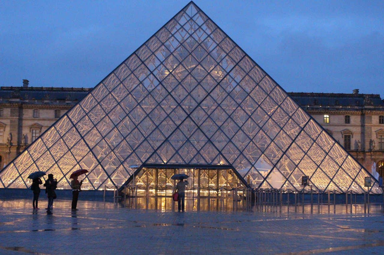 A glass pyramid of the Louvre Museum in the centre of a large square light up at night fall in Paris