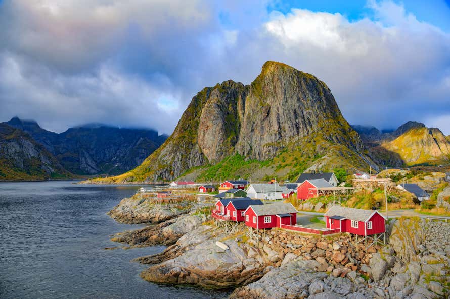 The Norwegian town of Reine in the Lofoten Islands. Red houses on rocky cliffs overlooking the sea with mountains in the background.