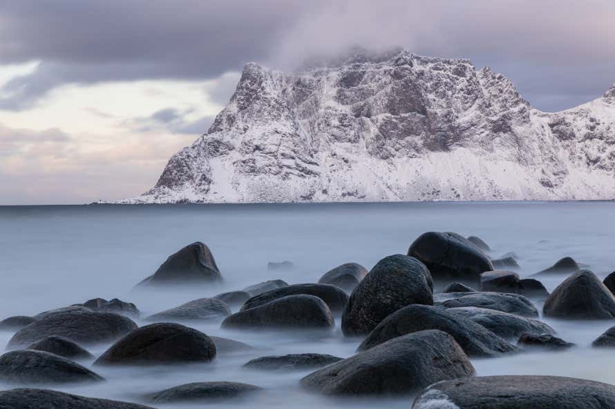 Uttakliev Beach in the Lofoten Islands with submerged rocks, mist, and snowy mountains in the background.
