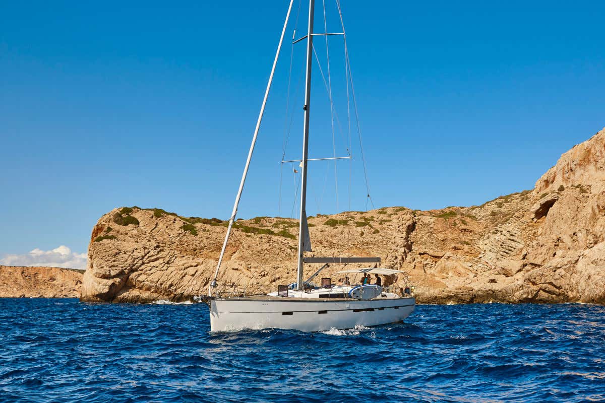 Un barco de vela de color blanco navegando frente a las costas de la isla de Cabrera en un día despejado.