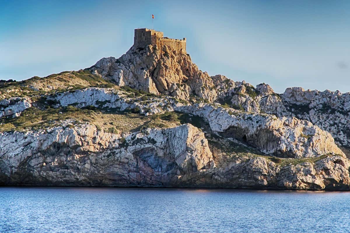 Mar en calma frente a los acantilados costeros de la isla de Cabrera coronados por una fortaleza con una bandera ondeando al viento.