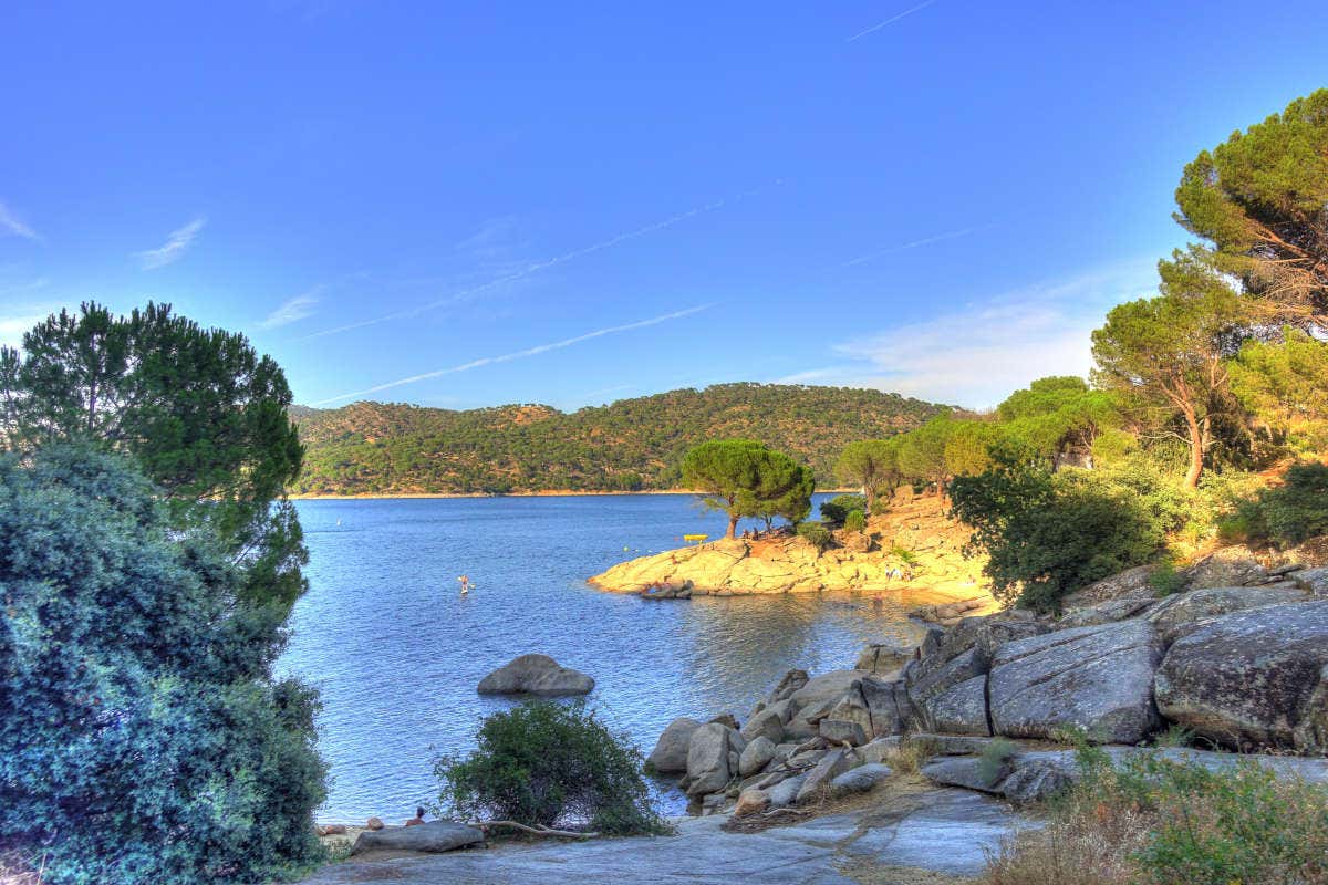 El pantano de San Juan rodeado de piedras y vegetación en un día despejado con una persona practicando paddle surf en sus aguas