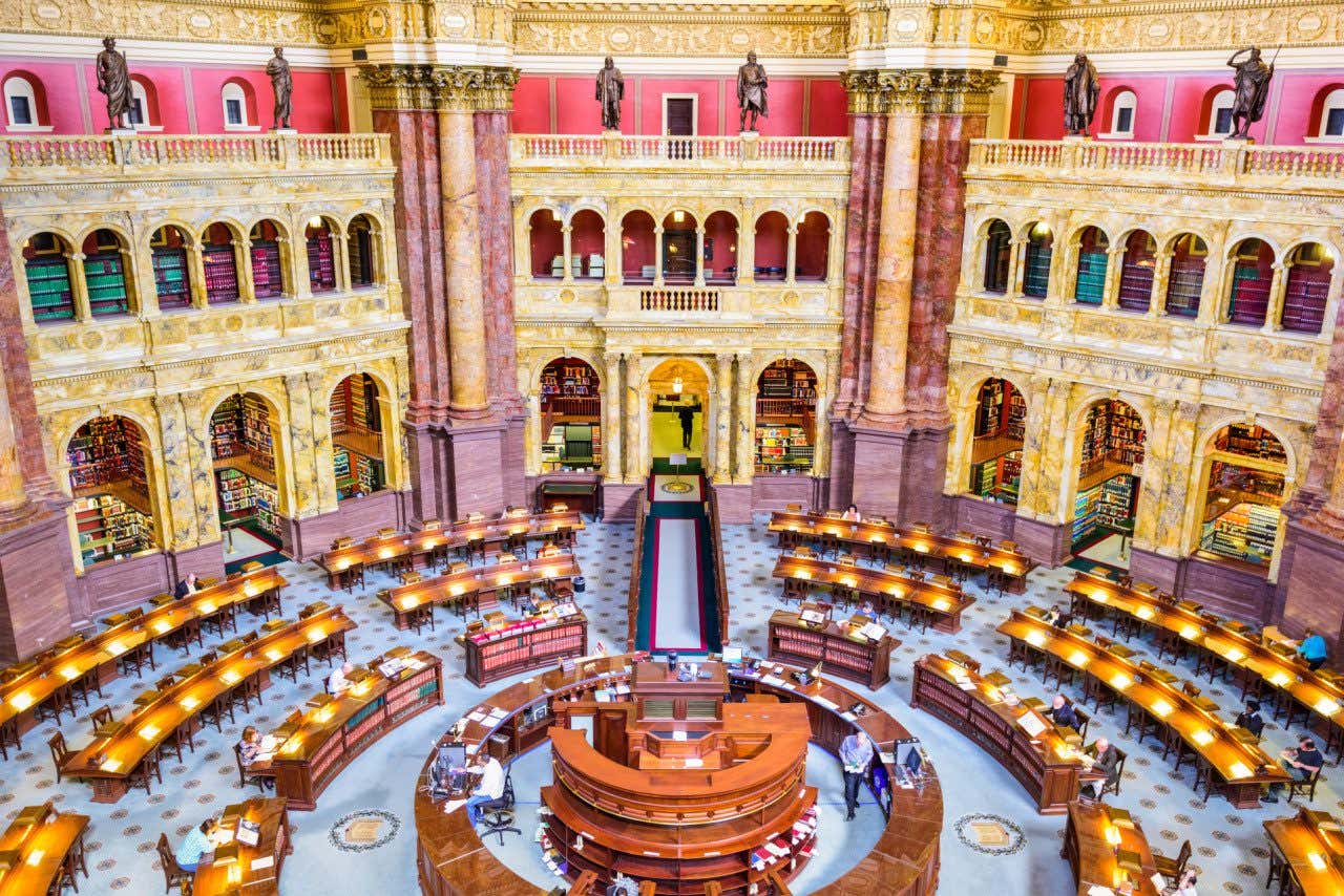 Interior da Biblioteca do Congresso dos Estados Unidos, em formato ovalado, com mesas iluminadas e alguns visitantes lendo