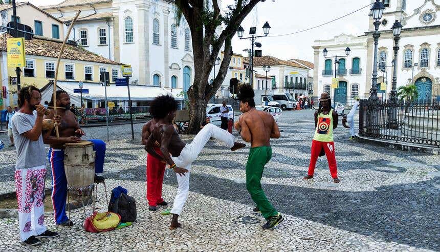 Dois homens jogando capoeira ao ar livre enquanto outros dois tocam os instrumentos