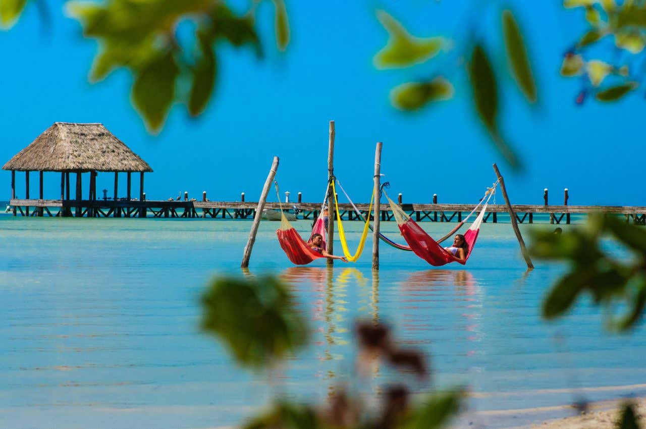 Two people relaxing in pink hammocks in shallow water with a wooden bridge behind them leading to a hut with a thatched roof