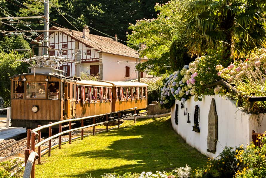 A two-carriage wooden tourist train in Sare passing a large white house with brown wooden beams.