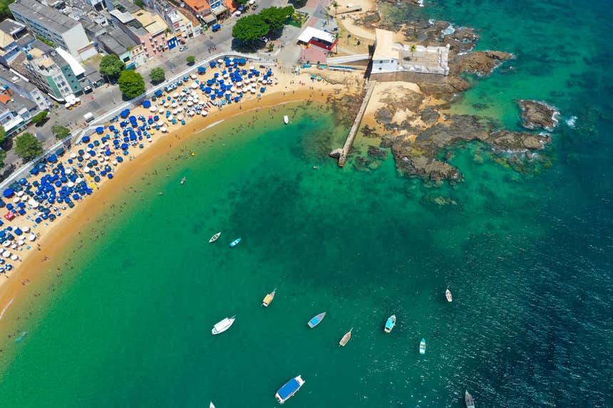 Vista aérea do Porto da Barra, com a areia repleta de guarda-sóis e alguns barcos nas águas verdes do mar