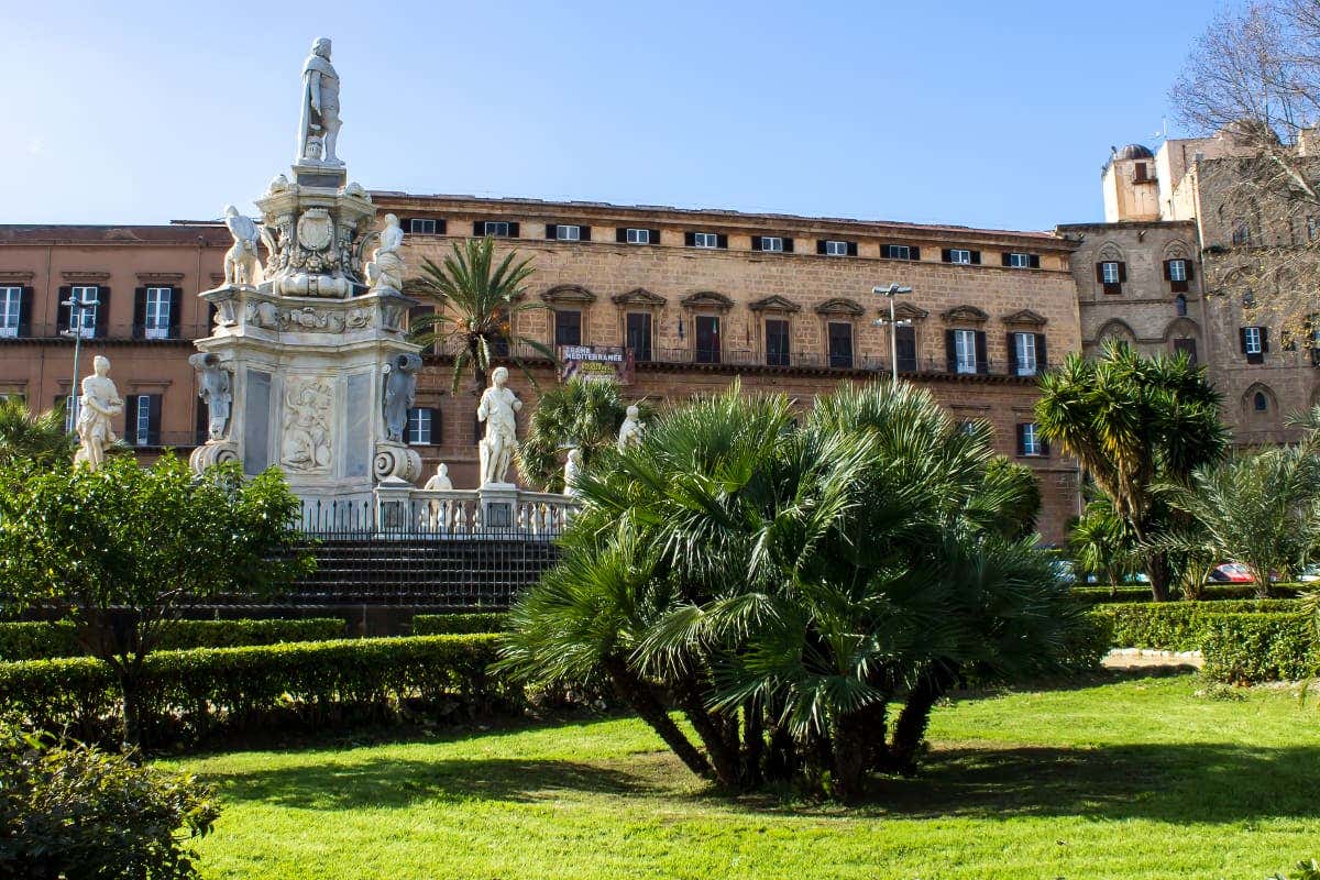 Il Palazzo dei Normanni, visto dall'esterno, con un grande monumento di statue bianche in un parco con felci e altri alberi