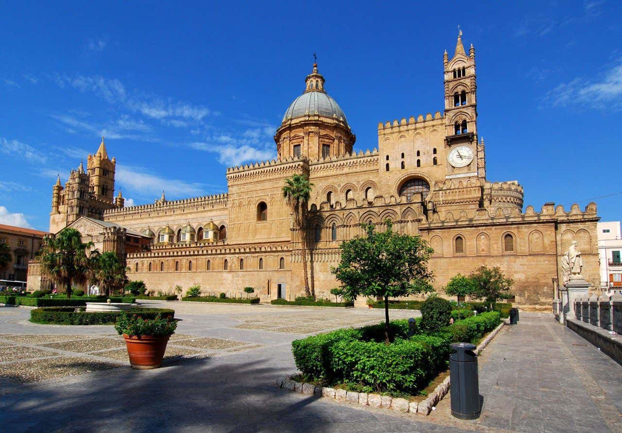 La Cattedrale di Palermo, un imponente edificio con torri e una cupola centrale e con di fronte un patio con alcune piante e alberi