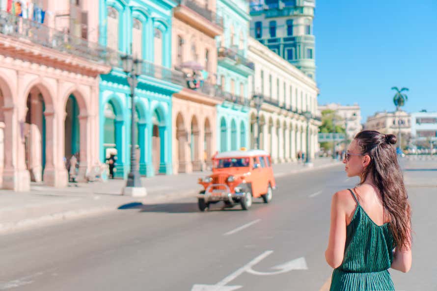 Ragazza di spalle che cammina per le strade di L'Avana davanti a un'auto e alle facciate colorate delle case