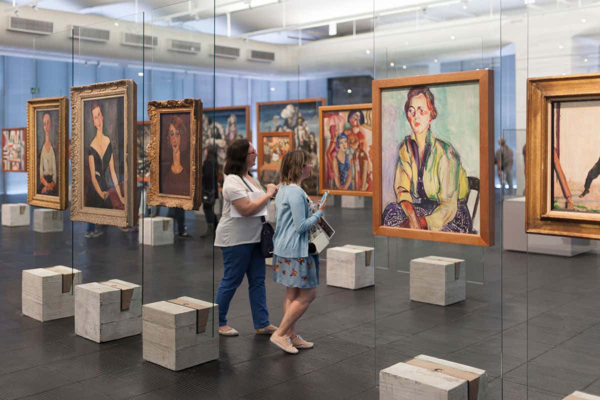 Duas visitantes com celular nas mãos apreciando obras do MASP. A maioria das pinturas exibe figuras de mulheres