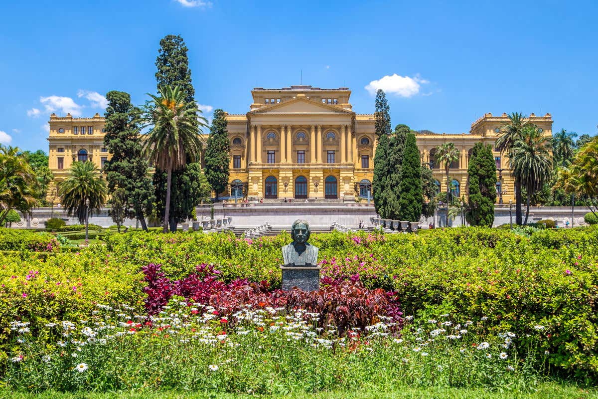 Belos jardins com flores, árvores e um busto de bronze em frente à fachada do Museu Paulista