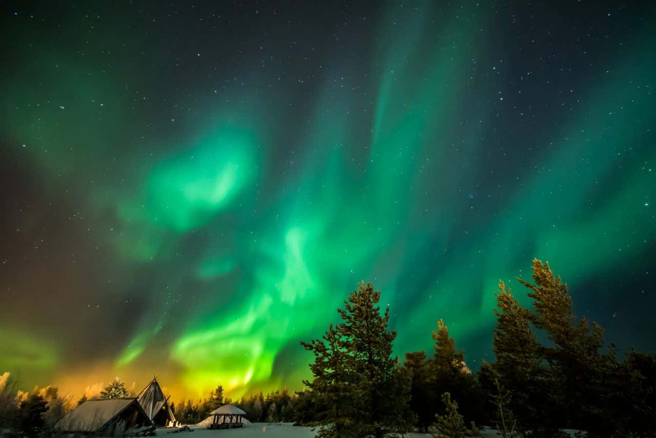 Intense colours of green, yellow and orange hues the Northern Lights in Rovaniemi with trees and dwellings