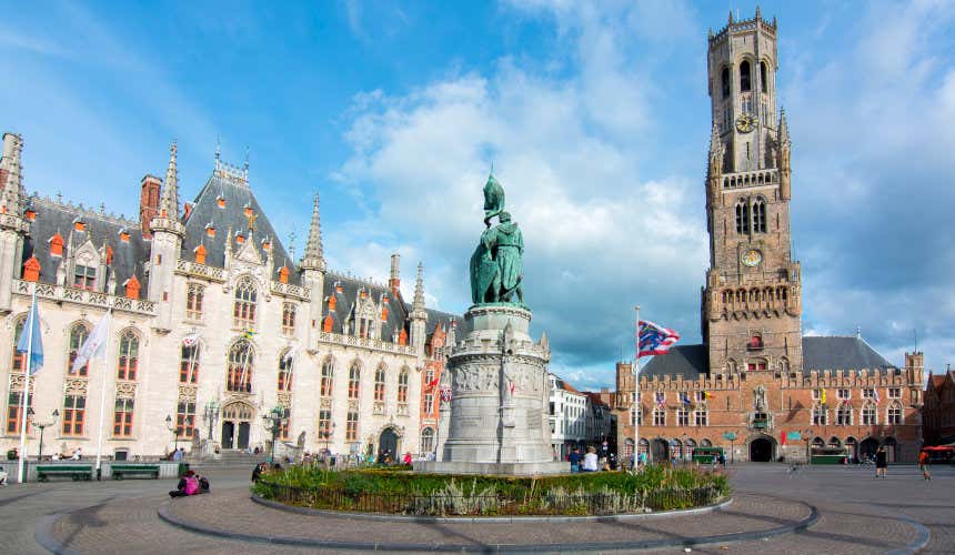 Vista sulla piazza Grote Markt a Bruges con Il campanile del Belfort e altri edifici ai suoi lati. Un'imponente statua si trova nel mezzo.