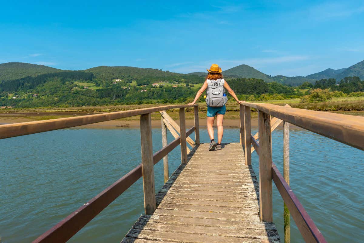 A woman wearing a hat and backpack with her back to the camera on a bridge admiring the water and vegetation of the Urdaibai Biosphere Reserve.