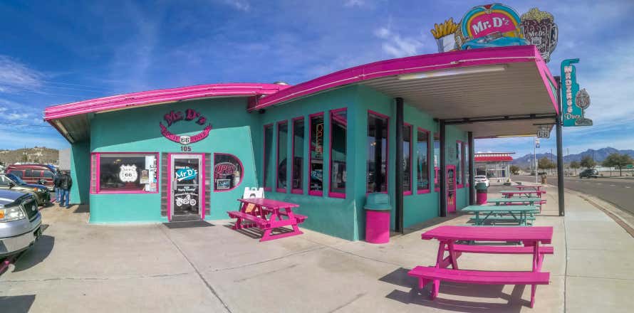 Tipico locale in stile anni '50, dai colori pastello, situato lungo la 66 route, con auto parcheggiate da un lato e tavolini all'aperto dall'altro