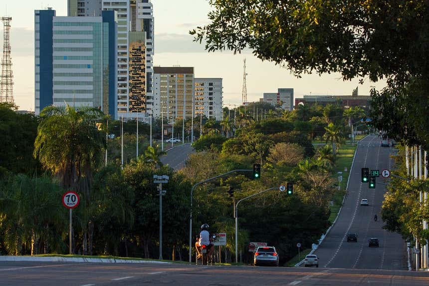 Moto e veículos circulam pela Avenida Joaquim Teotônio Segurado, com uma faixa de árvores no meio e edifícios ao fundo