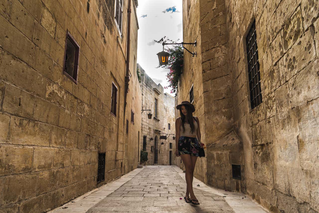 Una mujer joven vestida con un sobrero, falda y top, paseando por una estrecha calle de la ciudad de Mdina, en Malta